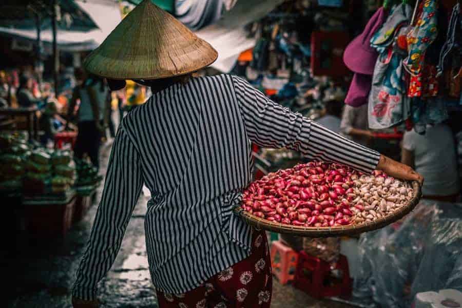 A woman, seen from behind carries a tray of onions in a market in VietnamCheapest Place in Southeast Asia